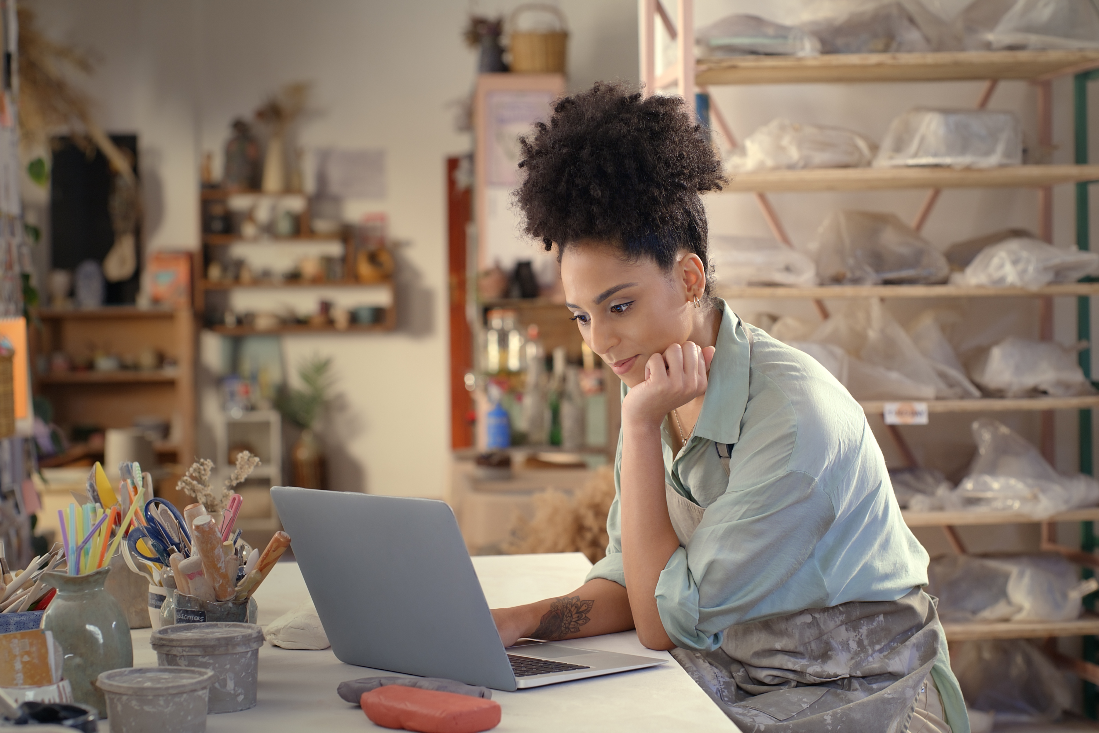 Crafting business owner using laptop in studio surrounded by wholesale craft supplies.