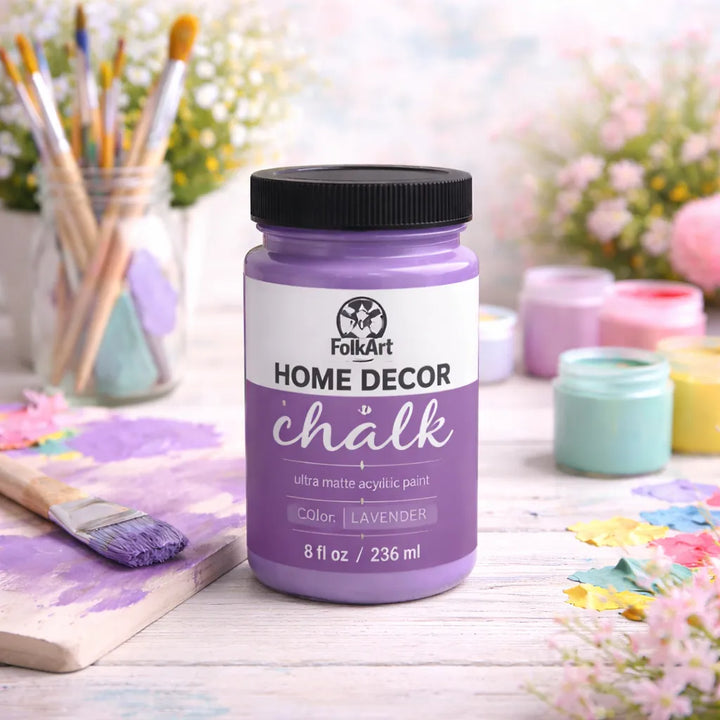 FolkArt Home Decor Chalk paint jar in lavender on a crafting table with brushes and flowers.